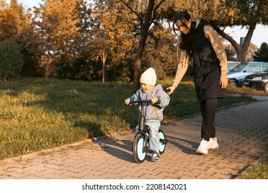 Happy Family Mother Teaches Child Son To Ride A Bike In The Park. Crying Boy Fear, Afraid Idea. Family, Childhood Concept. Mother Help Her Son Ride A Bicycle.