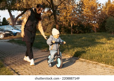 Happy Family Mother Teaches Child Son To Ride A Bike In The Park. Crying Boy Fear, Afraid Idea. Family, Childhood Concept. Mother Help Her Son Ride A Bicycle.