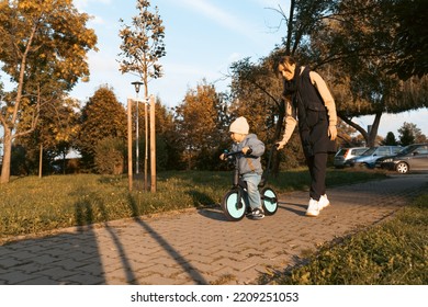 Happy Family Mother Teaches Child Son To Ride A Bike In The Park. Crying Boy Fear, Afraid Idea. Family, Childhood Concept. Mother Help Her Son Ride A Bicycle.