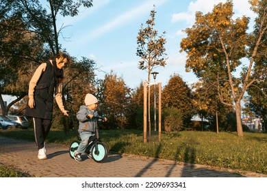 Happy Family Mother Teaches Child Son To Ride A Bike In The Park. Crying Boy Fear, Afraid Idea. Family, Childhood Concept. Mother Help Her Son Ride A Bicycle.