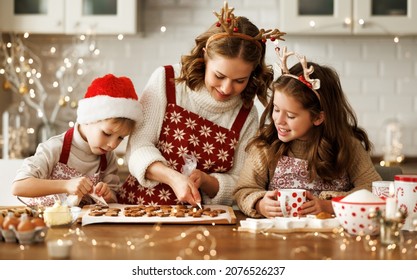 Happy Family Mother And Two Kids In Festive Outfit Making Christmas Cookies Together In Kitchen At Home During Winter Holidays, Caring Mom Decorating Xmas Gingerbreads With Small Children Siblings