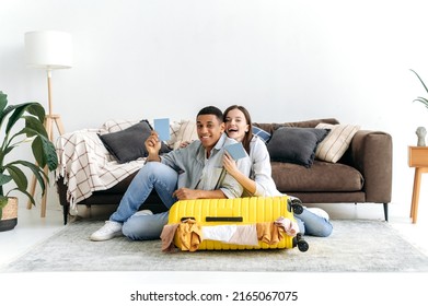 Happy Family Multiracial Couple, Mixed Race Guy And Caucasian Girl, Sit On The Floor At Home In The Living Room Near A Large Yellow Suitcase, Hold Passports, Prepare For A Long-awaited Vacation, Smile