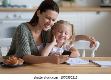 Happy Family Mum And Cute Little Kid Daughter Enjoy Lifestyle Hobbies Using Mobile App On Phone Look At Cellphone At Home, Smiling Mother With Child Girl Having Fun With Phone Sit At Kitchen Table