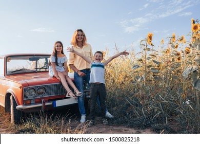 Happy Family Near Sunflowers Field, Mom With Two Children, Girl Teen In Dress Sitting On Hood Of The Red Retro Car, Mother And Son Standing Beside, Summer Vacation And Holidays