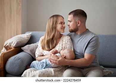 Happy Family With Newborn Baby. Parents Mom And Dad Hold Baby In Arms, Smile And Look At Each Other Sitting At Home On Couch