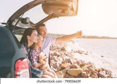 Happy Family On A Road Trip In Their Car. Dad, Mom And Daughter Are Traveling By The Sea Or The Ocean Or The River. Summer Ride By Automobile