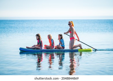 Happy Family On SUP Stand Up Paddle On Vacation. Active Family Riding SUP Boards And Paddling In The Ocean On A Beautiful Morning. Mother And Three Girls Share One SUP Board.