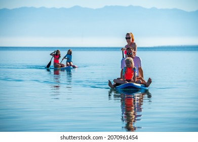 Happy Family On SUP Stand Up Paddle On Vacation. Active Family Riding SUP Boards And Paddling In The Ocean On A Beautiful Morning. Mother And Three Girls Share One SUP Board With Yachts In Background