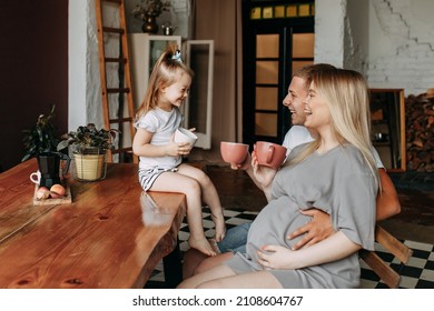 A Happy Family With One Child Having Breakfast Laughing Sitting At The Dining Table In A Loft-style Kitchen In A Cozy House. Pregnant Mom Dad And Little Daughter Cook Together. Selective Focus