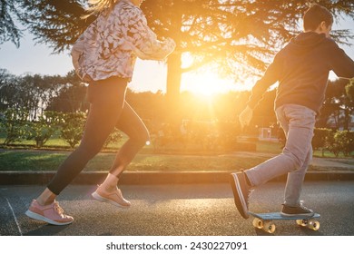 Happy family outdoors, mother and son go in sports, Boy rides skateboard, mom runs on sunny day. Silhouette people at sunset. Health care, authenticity, sense of balance and calmness.