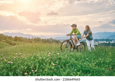 Happy Family Outdoors, Mother Teaching Son To Ride Bicycle. Cute Boy In Helmet Learn To Riding A Bike In Park On Green Meadow In Summer Day At Sunset Time.