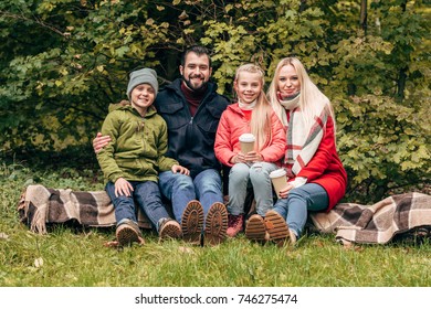 Happy Family With Paper Cups Smiling At Camera While Sitting Together In Autumn Park