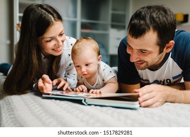 Happy Family, Parenthood And People Concept - Mother, Father With Baby Lying In Bed At Home. Portrait Of Young Smiling Family With Son Reading Book.