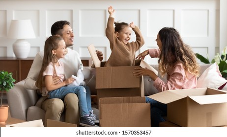 Happy Family Parents And Children Tenants Renters New Home Owners Playing On Moving Day Unpacking Boxes, Cute Little Child Daughter Jump Out Of Box Enjoying Relocation Into Own House Concept