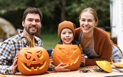 Happy Family Parents And Little Boy Son Carving Pumpkins In Backyard, Mother, Father And Child Smiling At Camera While Preparing Decorations For Halloween Outdoors, Making Jack-o-lantern Together