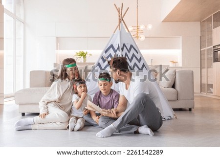 Happy family of parents and two children playing indian at home, wigwam tent, feather roach, reading book about native american indian culture