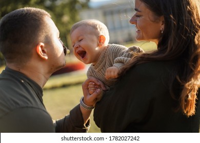 Happy Family In A Park. Dad Is Holding Dummy In Mouth. Mother Holding Baby Son Trying To Take Out A Pacifier