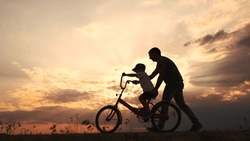 Happy Family In The Park. Father Teaching Son To Ride A Bike At Sunset Silhouette In The Park. Son Child Learning To Ride A Bike At Sunset Father Helping Son. Child Playing Lifestyle Riding Bike
