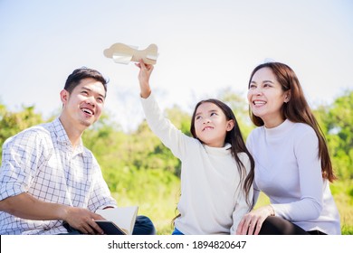 Happy Family Picnic. Asian Parents (Father, Mother) And Daughter Playing The Toy Airplane And Have Enjoyed Ourselves Together While Picnicking On Picnic Cloth In Green Garden In The Sunshine Day
