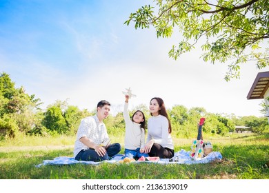 Happy Family Picnic. Asian Parents (Father, Mother) And Daughter Playing The Toy Airplane And Have Enjoyed Ourselves Together While Picnicking On Picnic Cloth In Green Garden In The Sunshine Day
