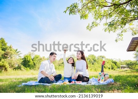 Happy family picnic. Asian parents (Father, Mother) and daughter playing the toy airplane and have enjoyed ourselves together while picnicking on picnic cloth in green garden in the sunshine day
