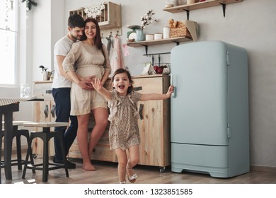 Happy Family. Playful Female Child Have Fun By Running In The Kitchen At Daytime Of Front Of Her Mother And Father.