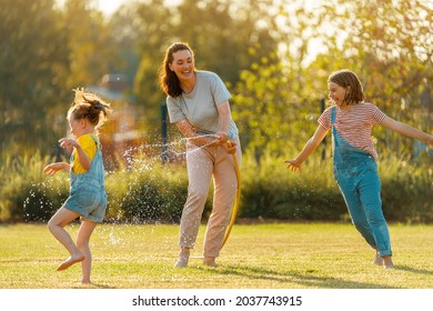 Happy Family Playing In Backyard. Mother Sprinkling Her Kids In Hot Summer Day.