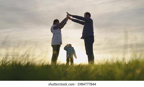 Happy Family Playing With Little Daughter, Family Symbol At Home In Sun. Mom, Dad And They Depict With Their Hands Roof Of House Over Head Of Child. Dream Of A Shared Home. Home Builders