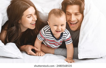 A happy family is playing peekaboo under a white blanket in bed. The parents are smiling and looking at their baby, who is laughing and reaching for the camera. - Powered by Shutterstock