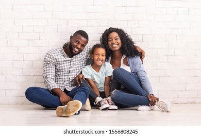 Happy Family Portrait. Cheerful african american parents and their little daughter sitting on floor and smiling together, white brick wall background.