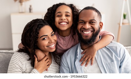 Happy Family. Portrait Of Cheerful African American Daughter Hugging Her Smiling Parents From Behind. Positive Woman, Man And Girl Posing For Photo And Looking At Camera At Home, Sitting On Sofa