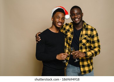 Happy Family Portrait On Christmas, Gay Male Couple On Beige Background. A Pair Of Black Men In Love Clink Glasses Of Champagne. Cheers. Celebrate Christmas And New Year