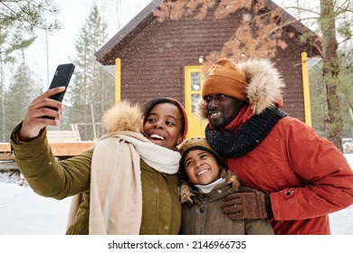 Happy family posing for selfie against country house on snowy winter day while young black woman with smartphone taking shot - Powered by Shutterstock