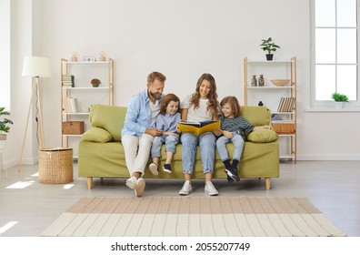 Happy Family Reading Book At Home. Mom, Dad, Little Kids Sitting Together On Green Sofa In New Big Light Living Room With White Walls, Beige Brown Rug, Wicker Storage Basket, And Shelves In Background