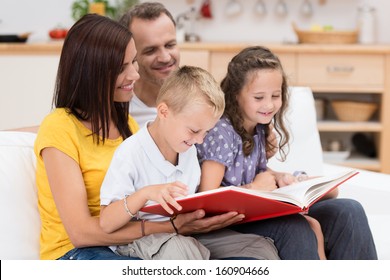 Happy Family Reading A Book Together With Attractive Young Parents Grouped With Their Two Small Children On A Couch In The Living Room