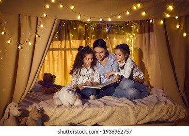Happy family reading good book in the evening. Young mother telling bedtime stories to little children. Mommy and daughters enjoying fairy tales sitting in cozy playroom tent decorated with LED lights - Powered by Shutterstock