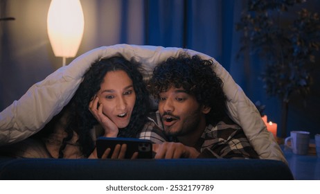 Happy family relaxed multiracial couple Indian man and Arabian woman at evening late night in home bed under blanket watching video movie film with mobile phone talking discussing emotional amazed - Powered by Shutterstock