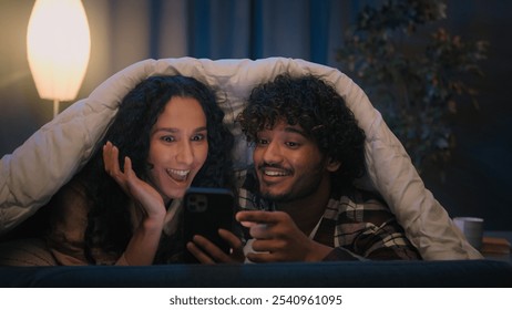 Happy family relaxed multiracial couple Indian man and Arabian woman at evening late night in home bed under blanket watching video movie film with mobile phone talking discussing emotional amazed - Powered by Shutterstock