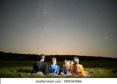 Happy family resting in night field looking at dark sky with many bright stars. Parents and children observing meteor shower.
