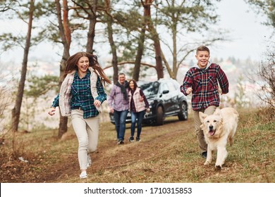 Happy Family Runs And Having Fun With Their Dog Near Modern Car Outdoors In Forest.