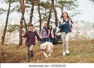 Happy Family Runs And Having Fun With Their Dog Near Modern Car Outdoors In Forest.