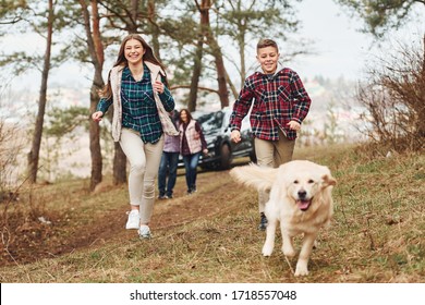 Happy Family Runs And Having Fun With Their Dog Near Modern Car Outdoors In Forest.