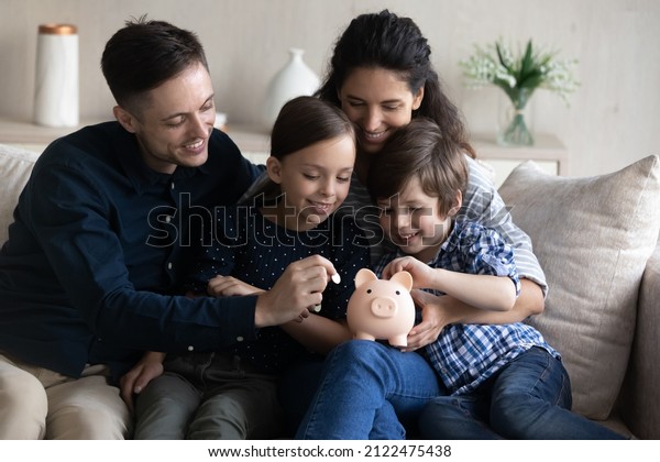 Happy family saving money for future purchase,\
making reserve fund, planning budget. Couple of parents and two\
little kids collecting cash, dropping coins into piggybank at home.\
Financial education
