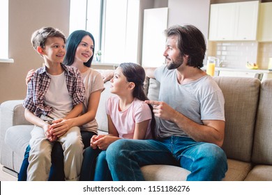 Happy family sits together on sofa. Boy is on mother's lap. Girl sits between woman and man. They are looking at each other.