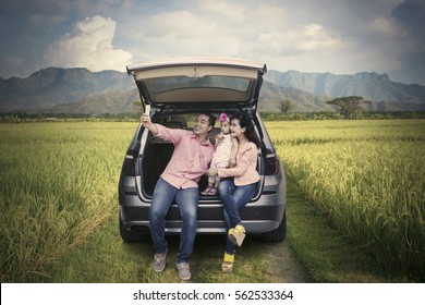 Happy Family Sitting Behind The Car While Taking Selfie Picture Using Smartphone In The Rice Field 