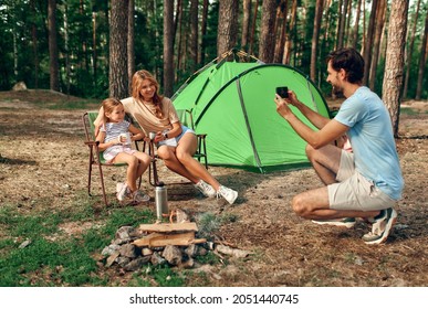 Happy Family Sitting By The Campfire Near The Tent In The Pine Forest For The Weekend. A Man Takes A Photo Of His Wife And Daughter On His Phone. Camping, Recreation, Hiking.