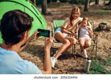 Happy Family Sitting By The Campfire Near The Tent In The Pine Forest For The Weekend. A Man Takes A Photo Of His Wife And Daughter On His Phone. Camping, Recreation, Hiking.