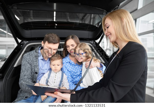 Happy\
family sitting in car trunk, buying new automobile in car\
dealership. Beautiful woman, car dealer holding black folder with\
document. Man signing contract, purchasing\
vehicle.