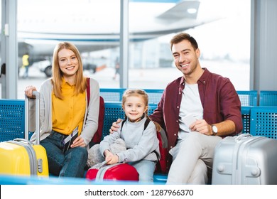 Happy Family Sitting In Departure Lounge And Smiling Near Luggage In Airport