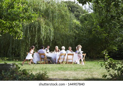 Happy Family Sitting At Dining Table While Girls Running Around In Garden